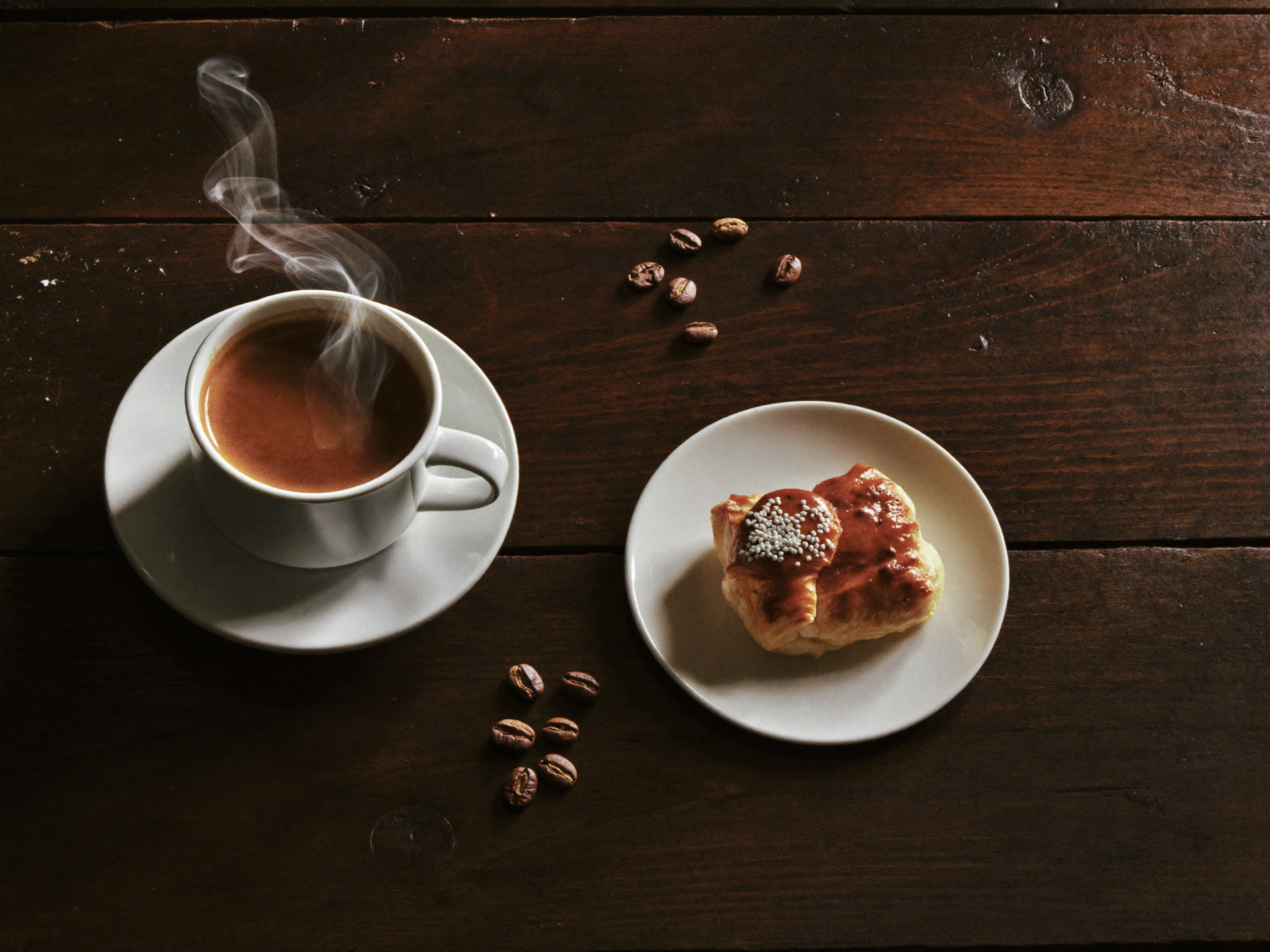 A pastry beside a steaming cup of coffee on a wood counter.
